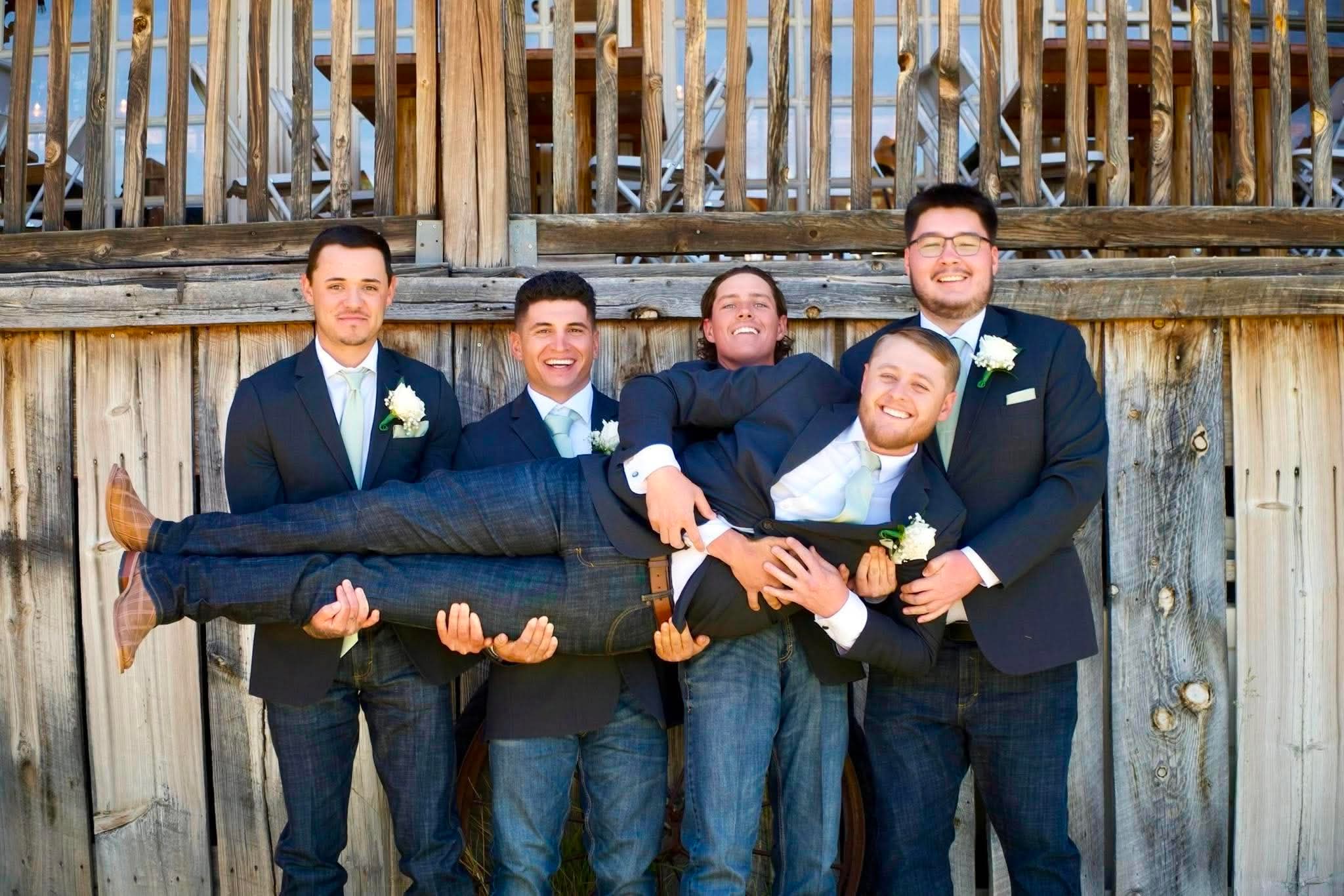 Groomsmen carrying the groom, laughing, in front of a barn